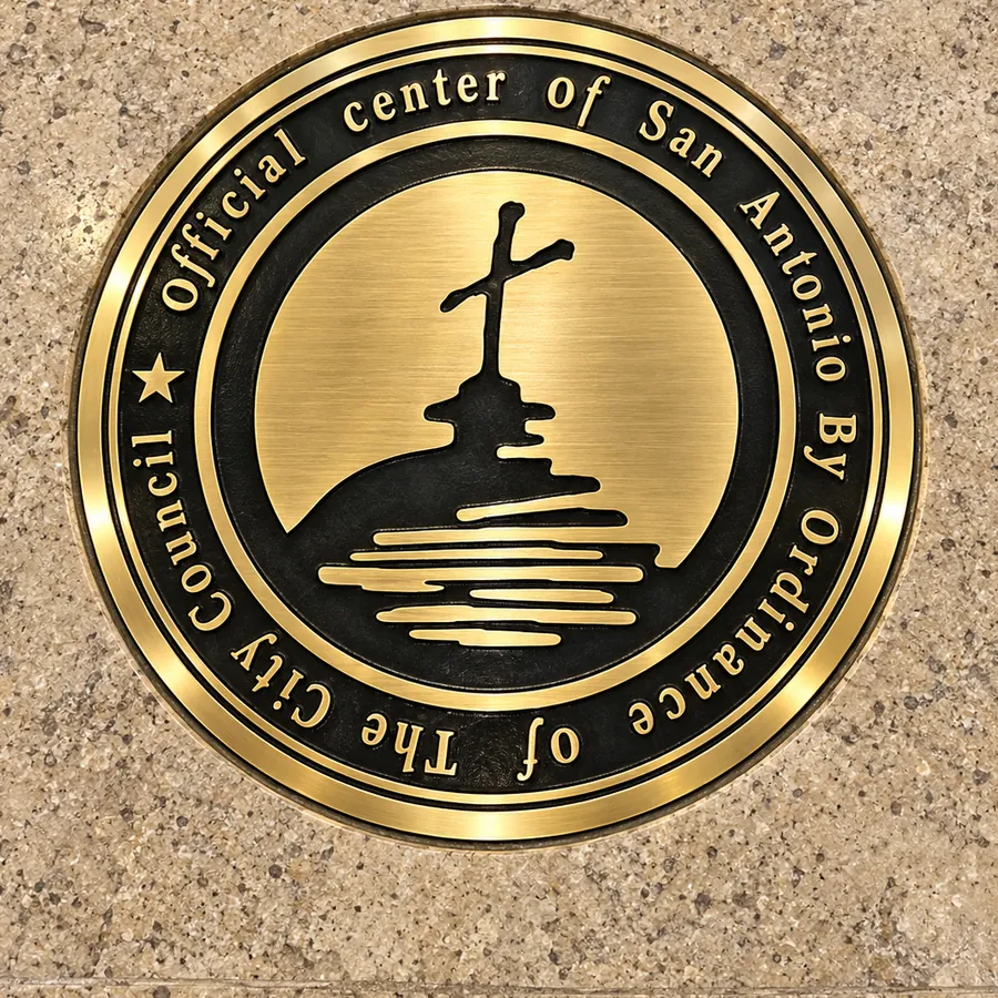 A bronze seal set into the stone floor of San Fernando Cathedral, reading: Official Center of San Antonio By Ordinance of The City Council, around a relief of a cross above the river.